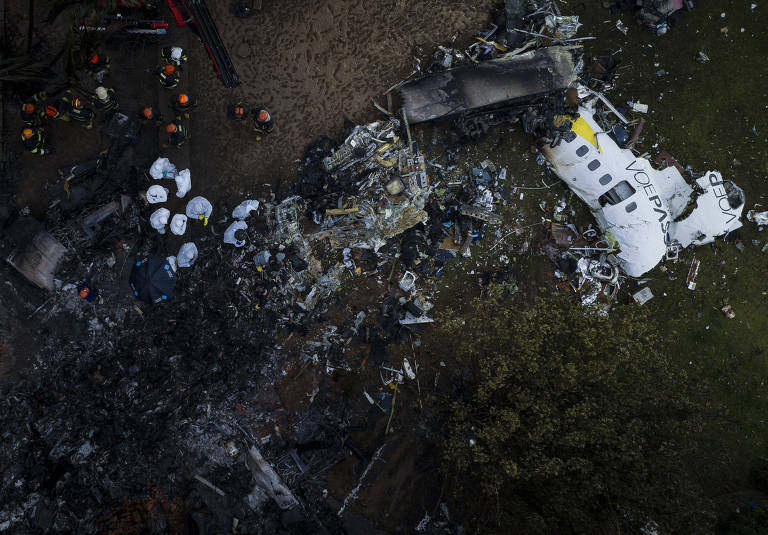 Imagem aérea de um acidente aéreo, mostrando destroços de uma aeronave em um terreno. Há equipes de resgate e socorro trabalhando na cena, com partes da fuselagem e outros destroços espalhados pelo chão.