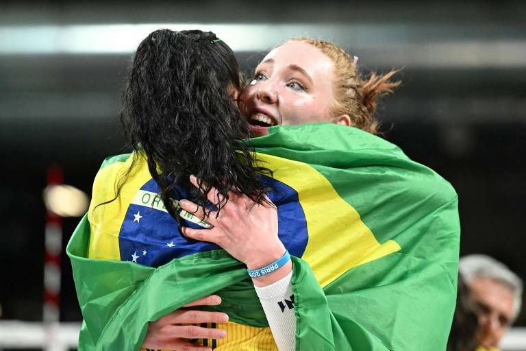 Julia Bergmann abraça colega, que está enrolada de costas e enrolada em uma bandeira do Brasil, na comemoração da conquista do bronze pela seleção de vôlei nas Olimpíadas de Paris-2024