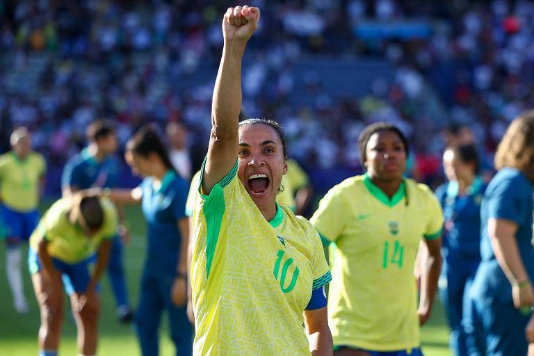 Uma jogadora de futebol, vestindo uma camisa amarela com detalhes verdes e um número 10, levanta o punho em sinal de celebração. Ela está sorrindo e parece estar muito animada. Ao fundo, outras jogadoras da equipe também estão presentes, algumas aplaudindo e outras em diferentes posturas, em um campo de futebol com uma multidão ao fundo.