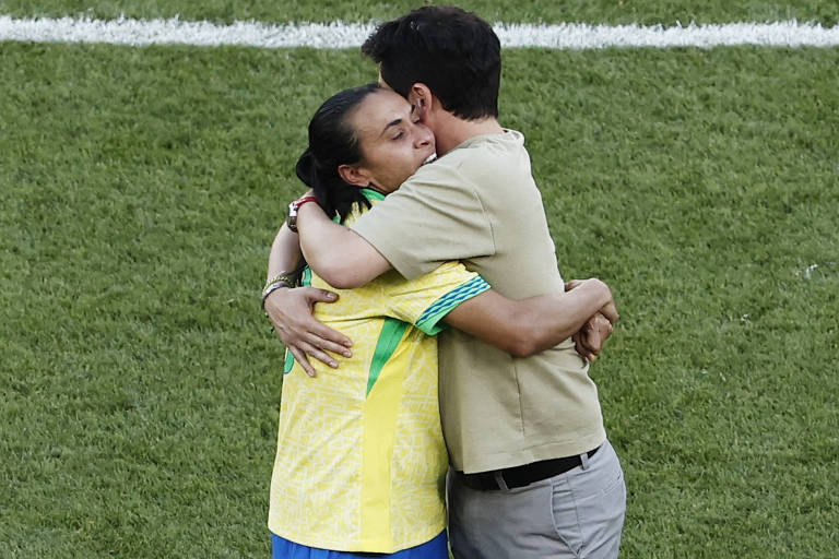 Uma mulher vestindo uma camisa amarela com detalhes verdes e azuis está abraçando um homem que usa uma camiseta bege e calças cinzas. Eles estão em um campo de futebol, com grama verde ao fundo e uma linha branca visível no chão.