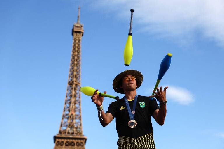 Usando chapéu de palha, Augusto Akio, o Japinha, medalhista de bronze no skate park em Paris-2024, exibe-se com malabares nas proximidades da Torre Eiffel