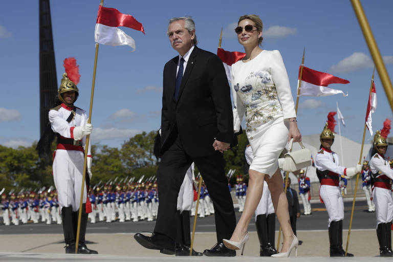 A imagem mostra um homem e uma mulher caminhando juntos em uma cerimônia oficial. O homem está vestido com um terno escuro e a mulher usa um vestido claro com detalhes. Ao fundo, há uma formação de soldados em uniforme, segurando bandeiras vermelhas e brancas. O céu está claro e há uma estrutura alta visível ao fundo.