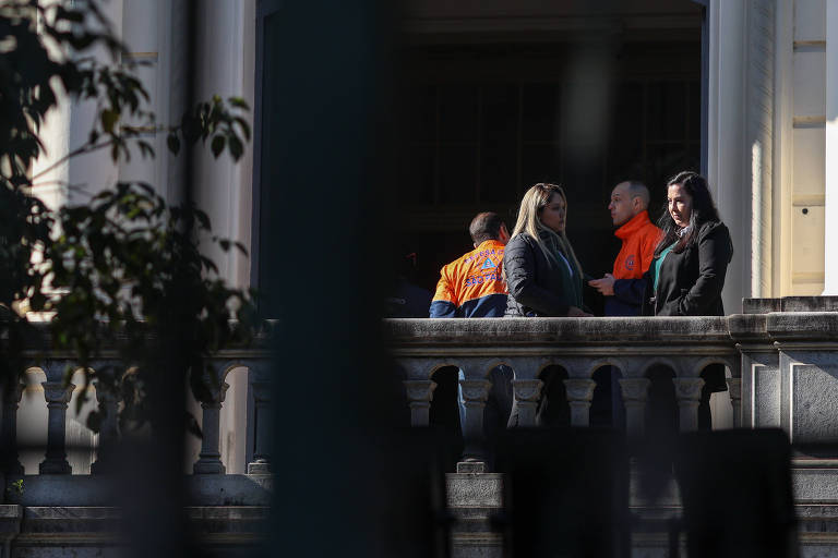 A imagem mostra um grupo de pessoas em uma conversa em um ambiente externo, em uma varanda. Algumas pessoas estão vestidas com uniformes laranja, enquanto outras usam roupas mais formais. A cena é parcialmente obstruída por grades e plantas, criando um efeito de profundidade.