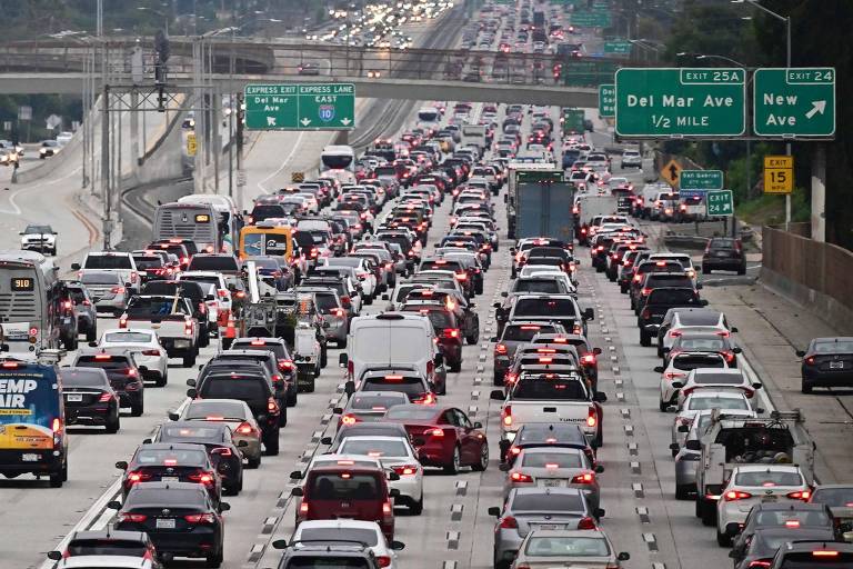 A imagem mostra uma rodovia com tráfego intenso, onde muitos carros estão parados ou se movendo lentamente. Há várias faixas de veículos, incluindo caminhões e carros de passeio. Ao fundo, é possível ver uma ponte e placas de sinalização. O céu está nublado, e a paisagem é urbana, com árvores e edifícios ao longe.