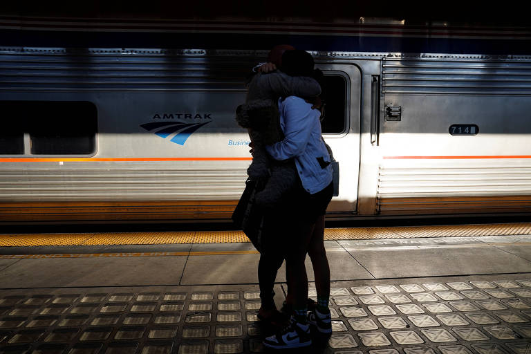 A imagem mostra duas pessoas se abraçando em uma estação de trem, com um trem em movimento ao fundo. A luz do sol cria um contraste, destacando as silhuetas das pessoas. O ambiente é urbano, com um piso de ladrilhos visível.