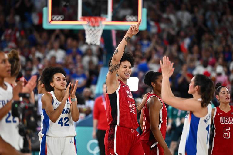 A imagem mostra jogadoras de basquete em um momento de celebração após uma partida. Uma atleta da equipe dos EUA, vestindo um uniforme vermelho com o número 15, levanta o braço em sinal de vitória, sorrindo. Ao fundo, outras jogadoras, incluindo uma da equipe da França com o número 42, aplaudem. O ambiente está cheio de torcedores, com uma cesta de basquete visível ao fundo.
