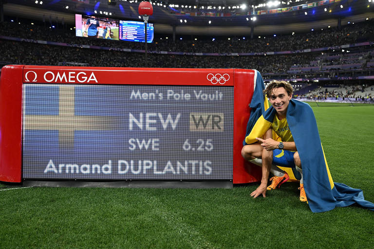 Sorrindo e agachado, o sueco Armand Duplantis posa ao lado de painel com o novo recorde mundial do salto com vara (6,25 metros), que ele registrou nas Olimpíadas de Paris