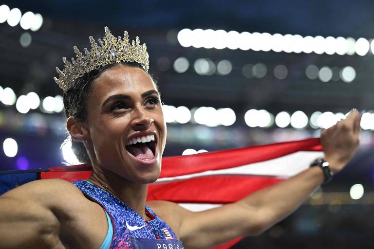 Com uma coroa na cabeça, sorrindo e segurando a bandeira dos EUA, Sydney Mclaughlin-Levrone festeja o ouro nos 400 m com barreiras nas Olimpíadas de Paris