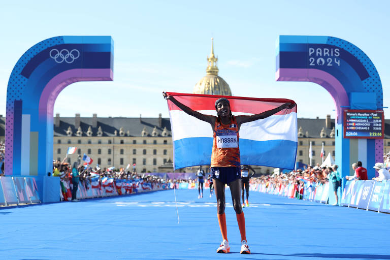 Um atleta está no final de uma corrida, segurando uma bandeira que combina as cores vermelho, branco e azul. O cenário é de um evento olímpico, com arcos de chegada ao fundo e um edifício histórico visível. O céu está claro e ensolarado.
