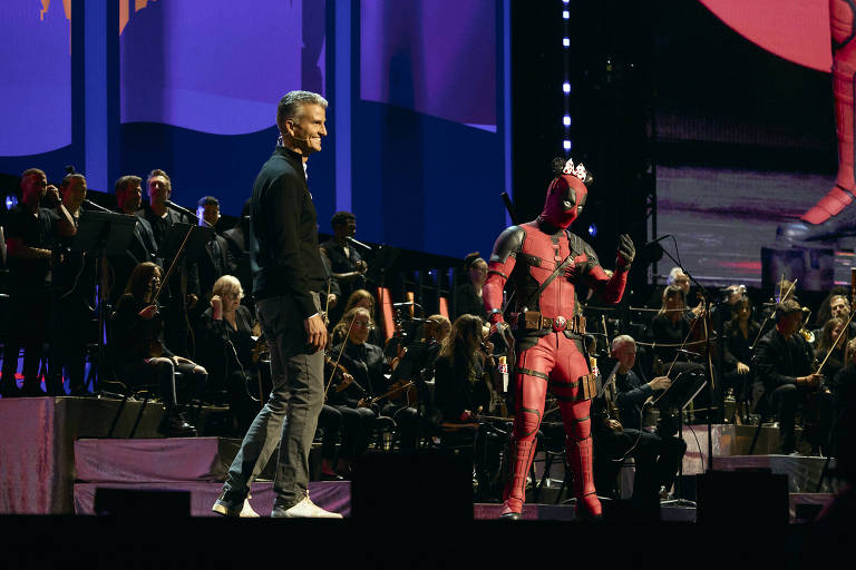 A imagem mostra um palco com duas figuras. À esquerda, um homem vestido com um traje casual, sorrindo e olhando para a direita. À direita, uma figura vestida com um traje vermelho e rosa, que parece ser uma fantasia de super-herói, fazendo uma pose. Ao fundo, há uma orquestra com músicos e uma iluminação colorida que destaca o ambiente.