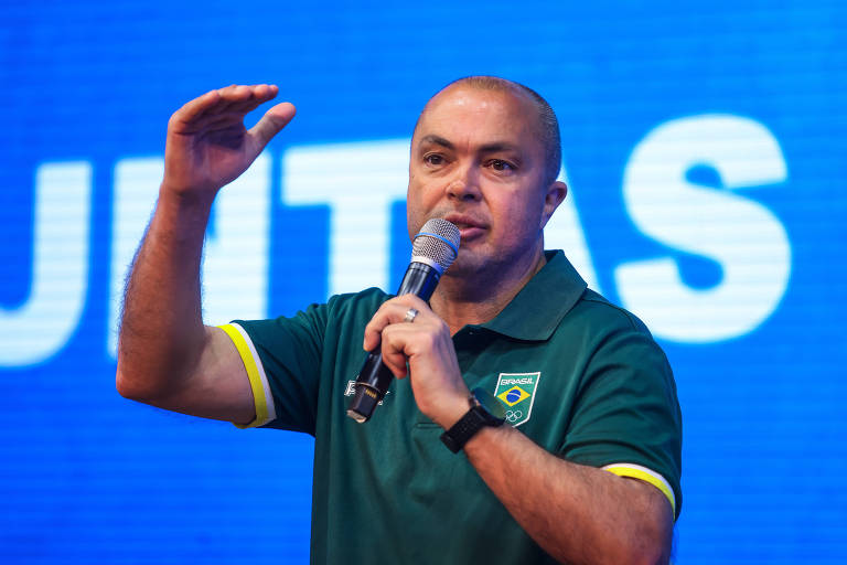 Um homem está em pé, segurando um microfone e gesticulando com a mão. Ele usa uma camiseta verde com detalhes amarelos, que remete às cores da bandeira do Brasil. Ao fundo, há uma tela azul com a palavra 'JUNTAS' em letras grandes e brancas.