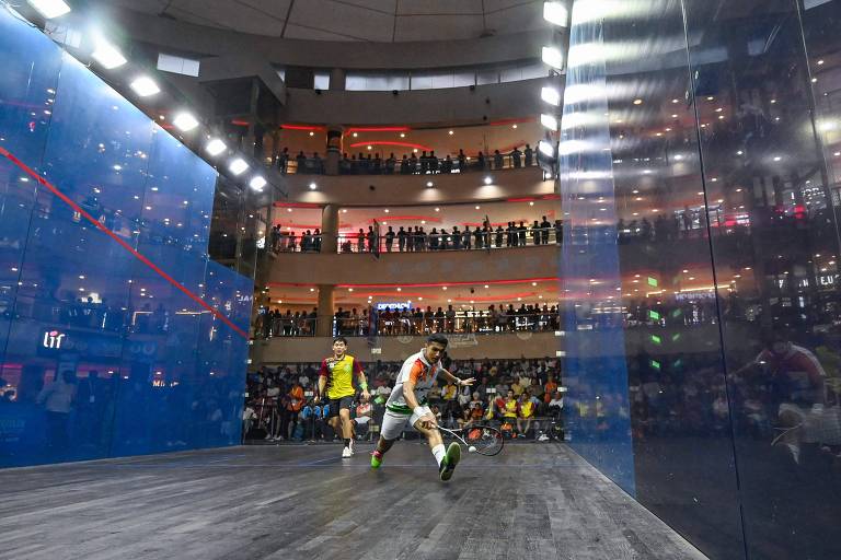 A imagem mostra uma partida de squash em um ambiente fechado, com dois jogadores em ação. Um jogador, vestido com uma camiseta branca e detalhes em verde e laranja, está se esticando para alcançar a bola, enquanto o outro, com uma camiseta amarela e vermelha, observa. Ao fundo, há uma plateia numerosa assistindo ao jogo, com várias pessoas em pé nas varandas do local. As paredes do campo de squash são de vidro azul, e há iluminação intensa no teto.
