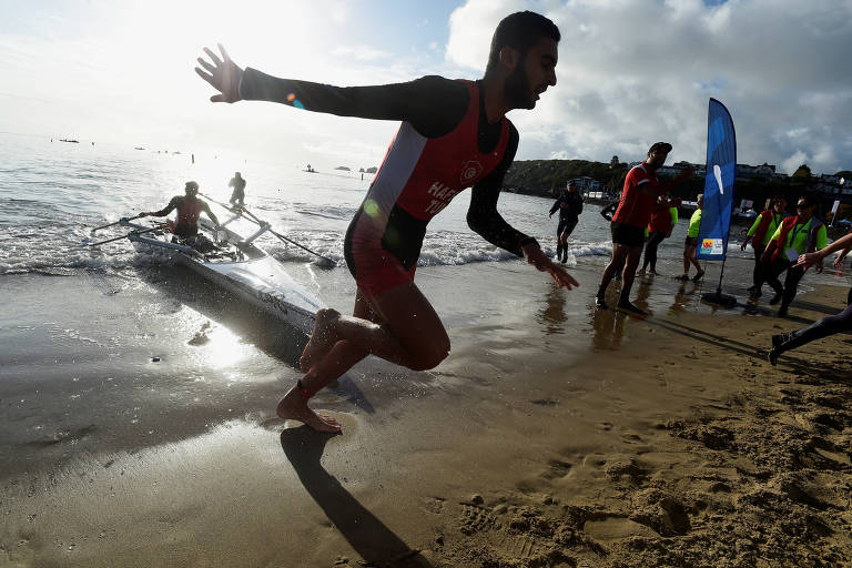 A imagem mostra um grupo de pessoas na praia durante uma competição de remo. Um homem em primeiro plano, vestindo um traje de competição vermelho, está correndo em direção à água, enquanto outros competidores estão ao fundo, alguns saindo da água e outros se preparando. Há um barco de remo parcialmente visível na água. O céu está parcialmente nublado, e a luz do sol reflete na superfície da água.
