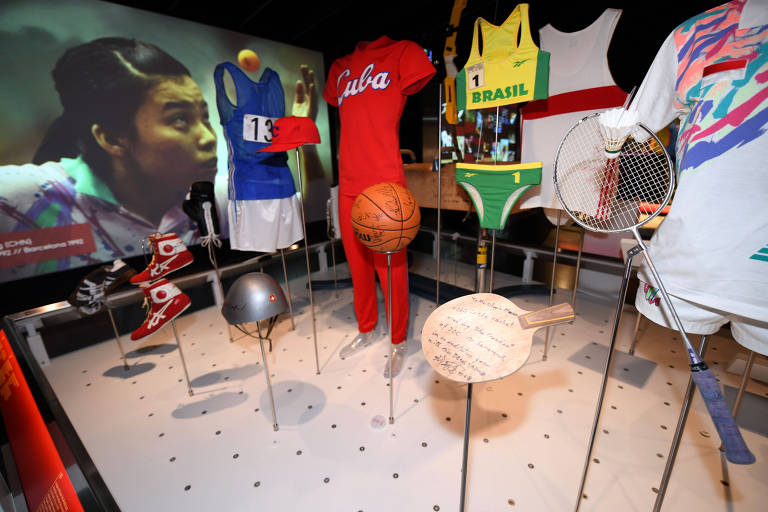 A imagem mostra uma exposição de roupas e equipamentos esportivos em um ambiente de museu. Há uma camiseta azul com o número 13, uma roupa vermelha com a palavra 'Cuba', um uniforme amarelo e verde do Brasil, um par de tênis de boxe, uma bola de basquete assinada, uma raquete de badminton e um capacete. Ao fundo, há uma projeção de uma atleta em ação, possivelmente durante os Jogos Olímpicos de Barcelona em 1992.
