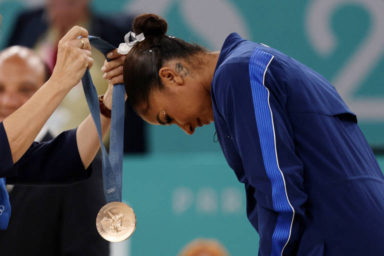 Uma atleta está recebendo uma medalha de bronze durante uma cerimônia olímpica. Ela está inclinando a cabeça para baixo enquanto uma mulher, que parece ser uma oficial, coloca a medalha ao redor de seu pescoço. A atleta está vestindo um uniforme azul e tem um coque preso com um laço. Ao fundo, há uma plateia desfocada.
