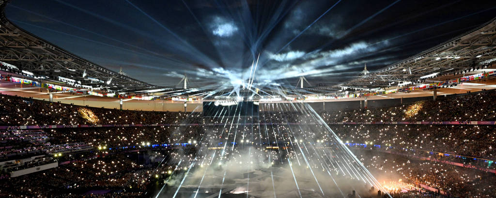 A imagem mostra um grande estádio iluminado por feixes de luz e efeitos de fumaça. O ambiente está repleto de espectadores, com uma atmosfera vibrante e festiva. O céu acima do estádio é visível, e as luzes criam um efeito dramático no espaço.
