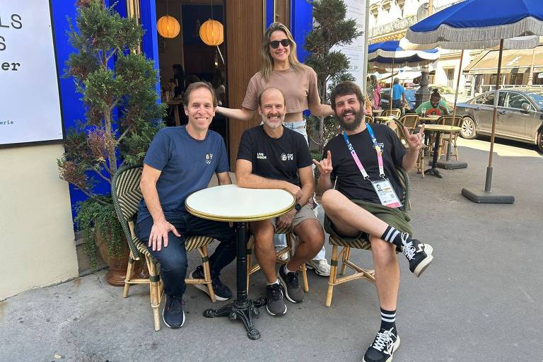 Quatro pessoas estão sentadas em um café ao ar livre. Três homens estão sentados à mesa, enquanto uma mulher está em pé atrás deles. O homem à esquerda usa uma camiseta azul e jeans, o do meio está com uma camiseta preta e shorts, e o da direita veste uma camiseta preta e shorts verdes. A mulher usa uma blusa clara e está sorrindo. Ao fundo, há mesas e cadeiras de café, além de uma árvore e uma placa azul com texto. O ambiente é ensolarado.