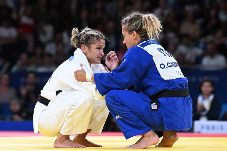 Duas atletas de judô estão em uma competição. Uma delas veste um judogi branco e a outra um judogi azul. Ambas estão em uma posição baixa, com as mãos se segurando, em um ambiente de competição com uma plateia ao fundo.