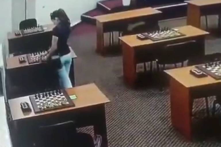 A imagem mostra uma sala com mesas dispostas em fileiras e tabuleiros de xadrez em cima de cada uma. Uma mulher está em pé ao lado de uma mesa, aparentemente jogando alguma coisa sobre ela. As mesas têm computadores e outros equipamentos. O ambiente é bem iluminado e possui um carpete escuro.