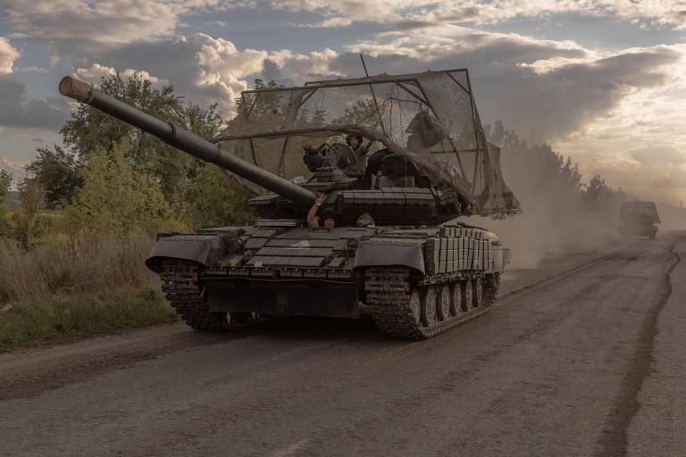 Numa estrada empoeirada, um tanque de guerra escuro, com uma grade sobre sua torre, segue com outro veículo atrás