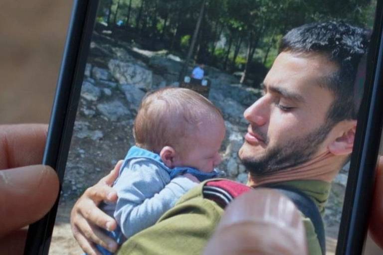 A imagem mostra um homem segurando um bebê em seus braços. O homem tem cabelo curto e está usando uma camiseta verde. O bebê, que está vestido com um macacão azul, parece estar encostado no peito do homem. A foto foi tirada em um ambiente ao ar livre, com árvores ao fundo.