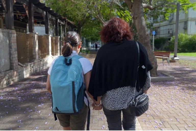 A imagem mostra duas pessoas caminhando de mãos dadas em um caminho de paralelepípedos. A pessoa à esquerda é uma criança com uma mochila azul clara, vestindo uma camiseta branca e shorts. A pessoa à direita é um adulto com cabelo cacheado e escuro, usando uma blusa preta e uma bolsa cruzada. O ambiente é um parque com árvores e flores caídas no chão.