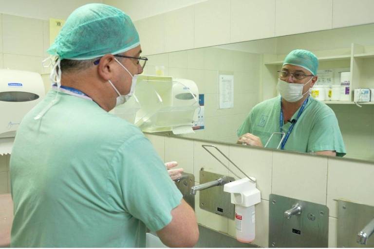 Um médico está de pé em frente a um espelho em um ambiente hospitalar. Ele usa um gorro cirúrgico verde e uma máscara facial. O médico está vestido com um uniforme cirúrgico verde e parece estar se preparando para um procedimento. Ao fundo, há equipamentos médicos e um dispenser de desinfetante para as mãos.