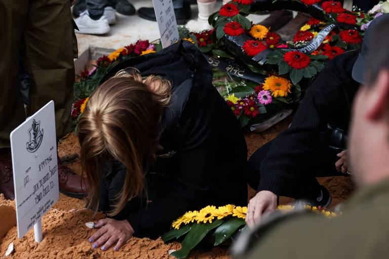 A imagem mostra uma cerimônia de sepultamento, onde uma mulher de cabelo longo e solto está ajoelhada sobre a terra, com as mãos apoiadas no solo. Ao fundo, há pessoas em pé, algumas vestindo uniformes militares. O local está decorado com flores coloridas, e há placas brancas visíveis, indicando os nomes dos falecidos.