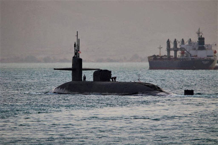 Silhueta de um submarino cinza-escuro em um mar cinzento. Ao fundo, um navio