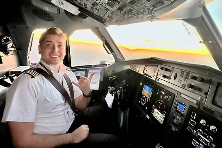 Um piloto sorridente está sentado no cockpit de um avião, com o painel de controle visível ao fundo. Ele usa uma camisa branca com insígnias de piloto e está fazendo um gesto de 'hang loose' com a mão. A luz do sol ilumina a cena, refletindo um pôr do sol através da janela do cockpit