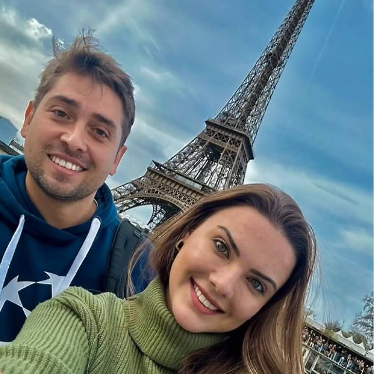 A imagem mostra um casal tirando uma selfie em frente à Torre Eiffel em Paris. O homem está à esquerda, sorrindo, vestindo um moletom azul com detalhes brancos. A mulher está à direita, sorrindo, usando um suéter verde. Ao fundo, a Torre Eiffel se ergue sob um céu nublado, e há pessoas e um lago visíveis na parte inferior da imagem