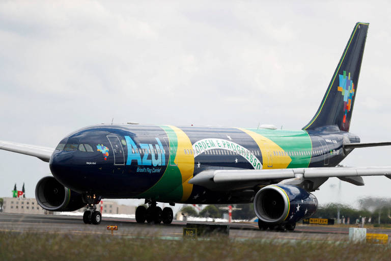 Um avião da companhia aérea Azul, decorado com as cores da bandeira do Brasil, está taxiando na pista. O fuselagem é predominantemente azul, com detalhes em verde e amarelo, e a palavra 'Azul' em destaque. A parte traseira do avião apresenta um design colorido. Ao fundo, é possível ver um aeroporto com bandeiras.
