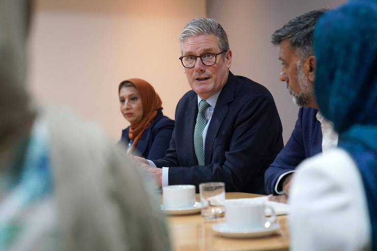 A imagem mostra uma reunião em uma mesa de conferência. Um homem de óculos e cabelo grisalho está falando, enquanto duas mulheres estão sentadas à sua esquerda e direita. A mulher à esquerda usa um hijab laranja, e a mulher à direita tem um hijab azul. Na mesa, há xícaras de café e copos de água.