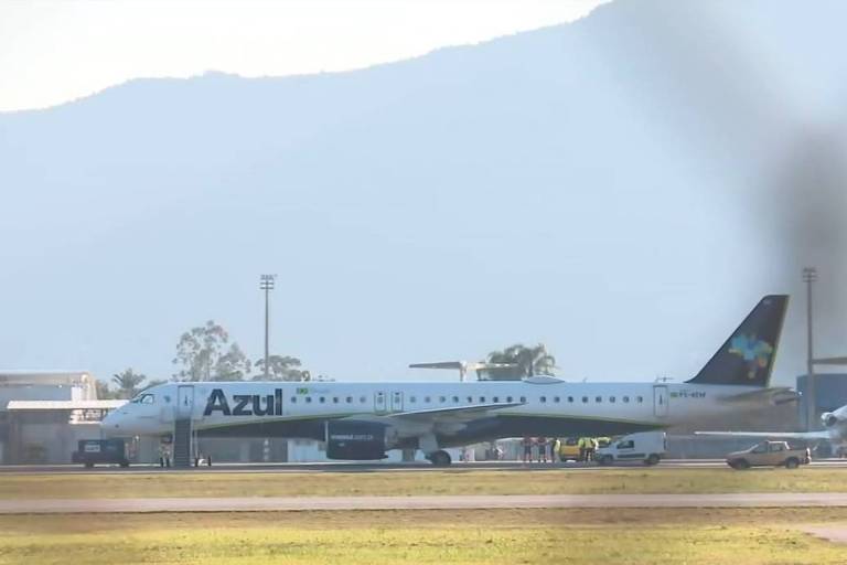 A imagem mostra um avião da companhia aérea Azul estacionado em um aeroporto. O avião é de fuselagem branca com a palavra 'Azul' em letras grandes e pretas. Ao fundo, há montanhas e um céu claro. Também são visíveis algumas estruturas do aeroporto e veículos de apoio ao lado do avião