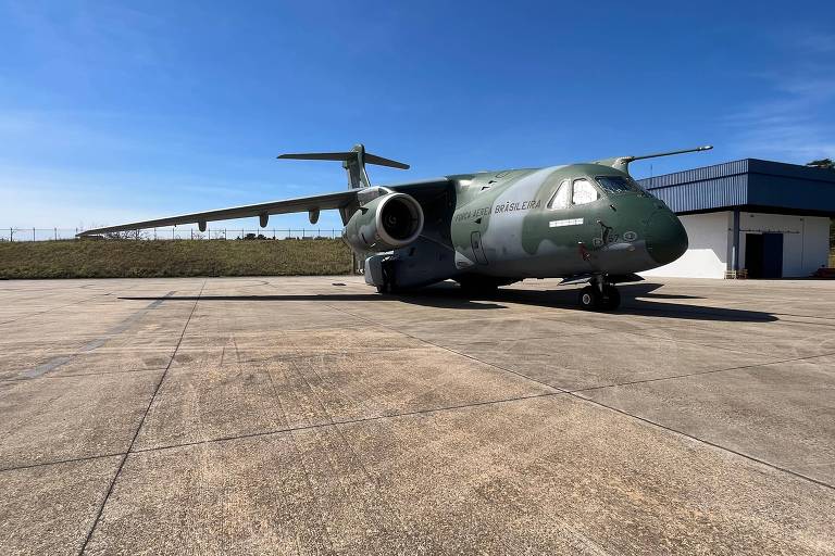 A imagem mostra um avião militar verde estacionado em uma pista de pouso. O céu está claro e azul, e ao fundo há um edifício de hangar. O avião possui uma fuselagem robusta e asas largas