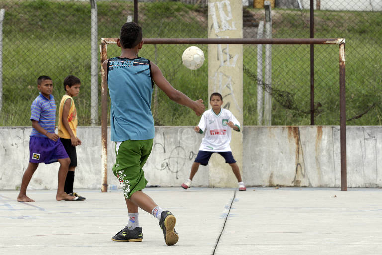 A imagem mostra um grupo de crianças jogando futebol em um campo. Um menino, de costas, está prestes a chutar a bola, enquanto outros três meninos observam. O goleiro, vestido com uma camiseta branca, está posicionado em frente ao gol. O campo é cercado por uma rede e há uma parede com grafites ao fundo.