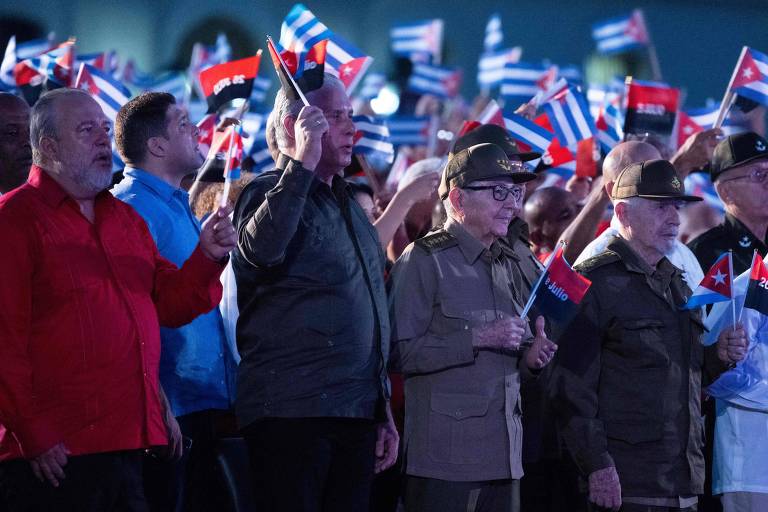 A imagem mostra um grupo de pessoas em um evento público, segurando bandeirinhas cubanas. Entre os presentes, há homens em trajes variados, incluindo um homem de camisa vermelha e outro de uniforme militar. Todos parecem estar em um momento de celebração ou homenagem, com expressões sérias e respeitosas