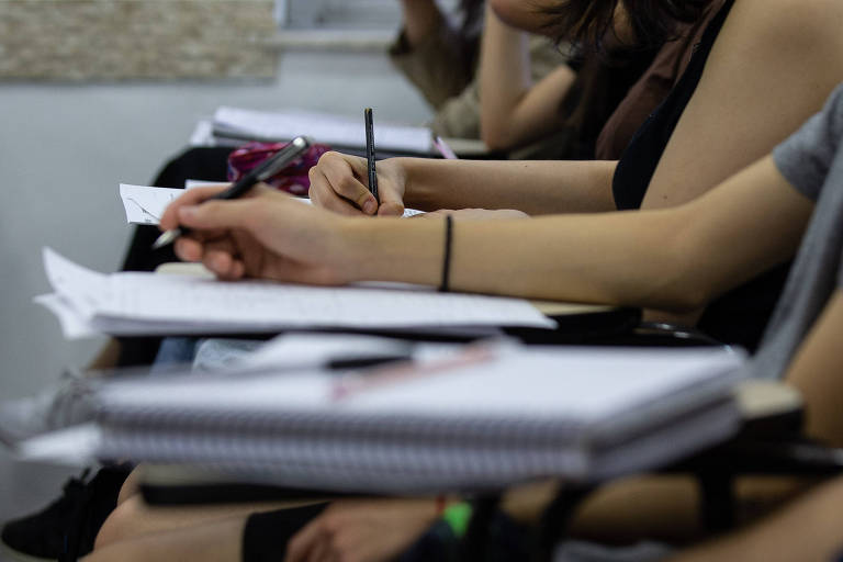 A imagem mostra um grupo de pessoas sentadas em uma sala de aula, focadas em suas anotações. As mãos de alguns participantes estão visíveis, segurando canetas e escrevendo em folhas de papel e cadernos. É possível ver uma variedade de roupas e estilos, com algumas pessoas usando camisetas e outras com blusas de manga. O ambiente parece ser um espaço educacional, com mesas e materiais de escrita.