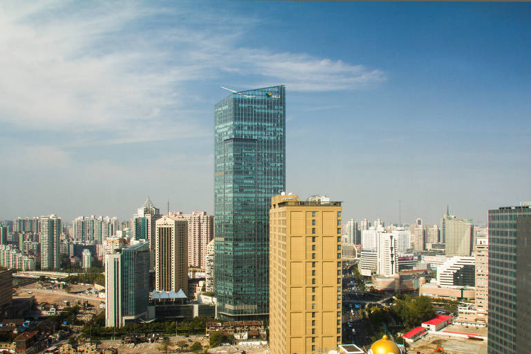 A imagem mostra uma vista panorâmica de uma cidade com vários edifícios altos. No centro, destaca-se um arranha-céu de vidro, refletindo a luz do sol. Ao redor, há outros prédios de diferentes tamanhos e estilos arquitetônicos, com um céu azul e algumas nuvens ao fundo