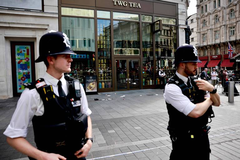 A imagem mostra dois policiais em uniforme, de pé em uma calçada, em frente a uma loja chamada TWG TEA. O ambiente é urbano, com edifícios ao fundo e pessoas visíveis através das janelas da loja. O dia parece ensolarado.