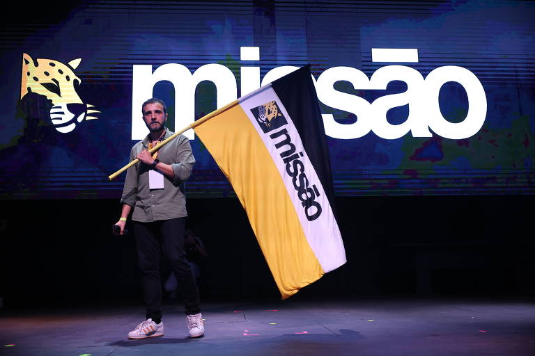 Um homem está em um palco segurando uma bandeira com as cores preta, amarela e branca, que possui a palavra 'missão' escrita. Ao fundo, há um grande painel com a mesma palavra 'missão' em letras brancas e um desenho de uma cabeça de jaguar à esquerda.