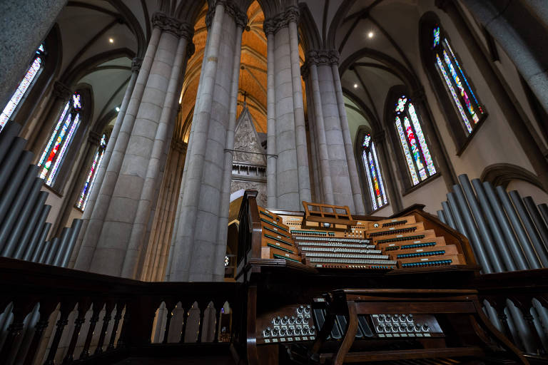 A imagem mostra o interior de uma igreja com um grande órgão em primeiro plano. O órgão é composto por várias teclas e tubos, e está situado em uma área com colunas altas e arcos. Ao fundo, há vitrais coloridos que filtram a luz, criando um ambiente iluminado. O teto é alto e apresenta detalhes arquitetônicos.