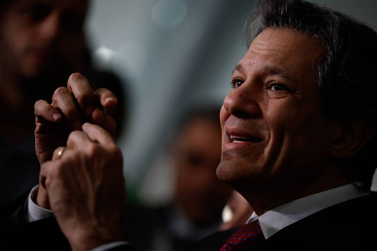 A imagem mostra Fernando Haddad, ministro da Fazenda, gesticulando com as mãos. Ele está em um ambiente com pouca luz, e outras pessoas são visíveis ao fundo, mas não são claramente identificáveis. O homem parece estar em um momento de conversa ou reflexão.