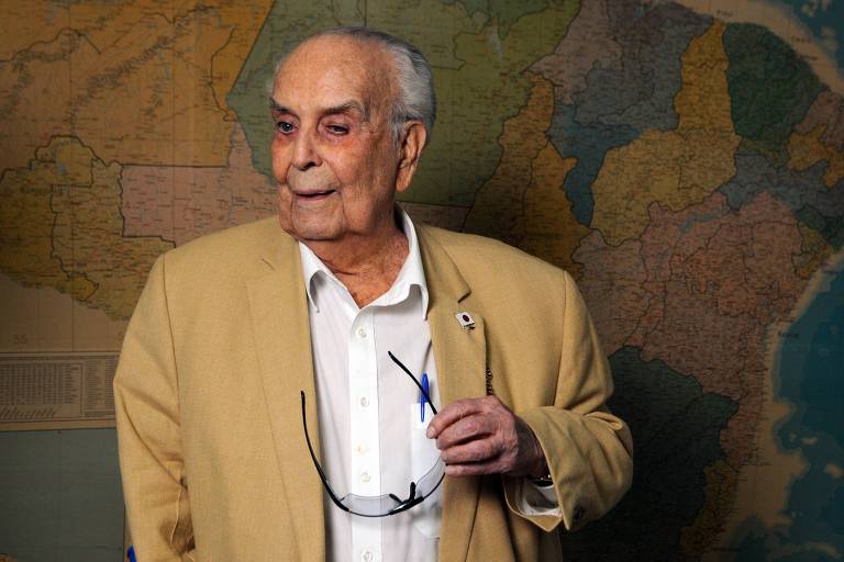 Um homem idoso está posando em frente a um mapa colorido. Ele usa um paletó claro e uma camisa branca, segurando óculos na mão. O fundo apresenta um mapa que mostra diferentes regiões, com cores variadas representando estados ou áreas geográficas