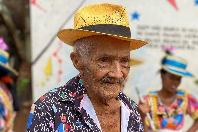 Idoso está vestido com camisa colorida e chapéu amarelo. Ele segura uma garrafa de refrigerante e parece estar em um ambiente festivo, possivelmente durante o Carnaval. Ao fundo, há pessoas vestidas com trajes de festa, com saias coloridas e chapéus, que também estão participando da celebração