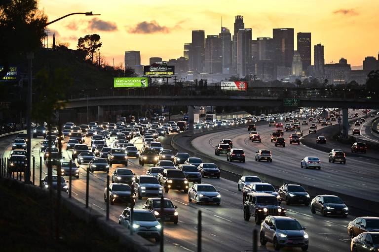 A imagem mostra uma vista do tráfego em uma rodovia de Los Angeles ao entardecer. Vários carros estão se movendo em direções opostas, com a cidade ao fundo, onde se destacam os arranha-céus. O céu apresenta tons de laranja e azul, indicando o pôr do sol. Há também painéis publicitários visíveis ao longo da rodovia