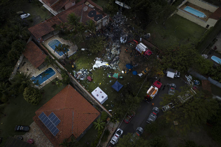 Imagem aérea de um local de acidente aéreo, mostrando destroços de uma aeronave em um quintal. Há várias viaturas de emergência, como ambulâncias e caminhões de bombeiros, além de pessoas e tendas no local. Ao fundo, pode-se ver uma piscina e várias casas na vizinhança.