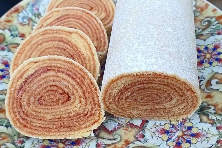 A imagem mostra um prato decorativo com fatias de bolo de rolo, que são camadas de massa enroladas com um recheio. O bolo está em um prato com um padrão floral, e ao lado há um rolo inteiro do bolo. Ao fundo, podem ser vistas plantas em vasos.