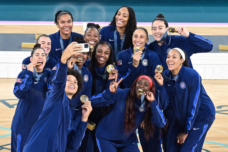 Um grupo de mulheres atletas, vestindo uniformes azuis, posam juntas para uma selfie, exibindo suas medalhas. Elas estão sorrindo e se divertindo, em um ambiente que parece ser uma quadra de basquete.
