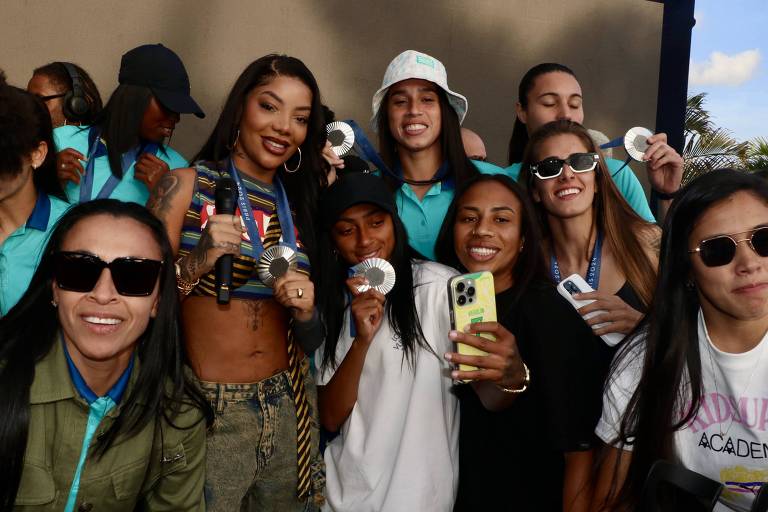 Um grupo de pessoas sorrindo em uma celebração, posando para a foto. No centro, uma mulher com cabelo longo e liso, usando uma blusa listrada, segura uma medalha. Ao redor dela, várias mulheres vestindo uniformes esportivos e outras roupas casuais, todas exibindo medalhas. Um homem à esquerda, com cabelo escuro e camisa azul, também sorri. 